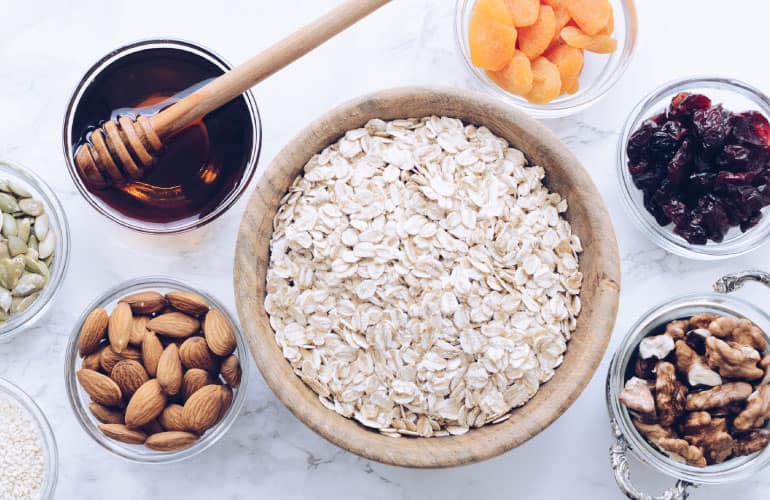 A dozen small glass bowls sitting on a marble countertop. The bowls contain ingredients such as raw almonds, pepitas, oats, walnuts, dried cranberries, coconut shavings, and dried apricots.